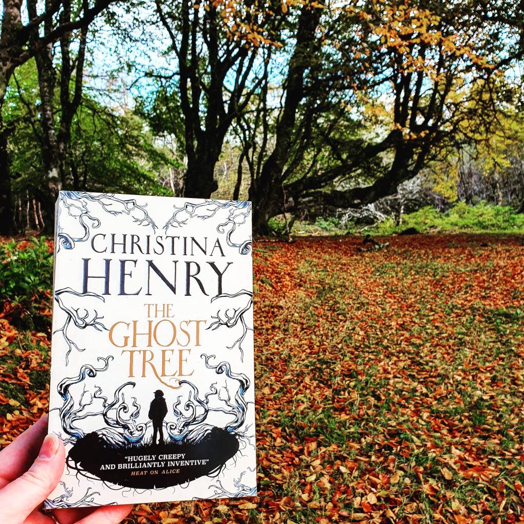 The Ghost Tree by Christina Henry (White cover with silhouettes) is held up against an autumnal forest scene.